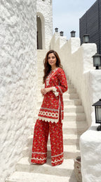 Woman in a red outfit with white patterns standing on white steps against a white wall.