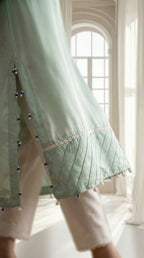 Light green top with decorative trim and white pants in a bright room.